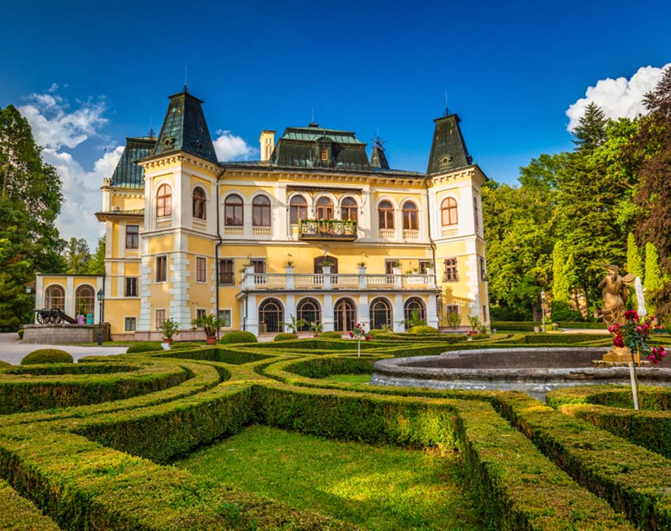 Historický kaštieľ obkolesený udržiavanou záhradou so sochou a fontánou počas slnečného dňa.