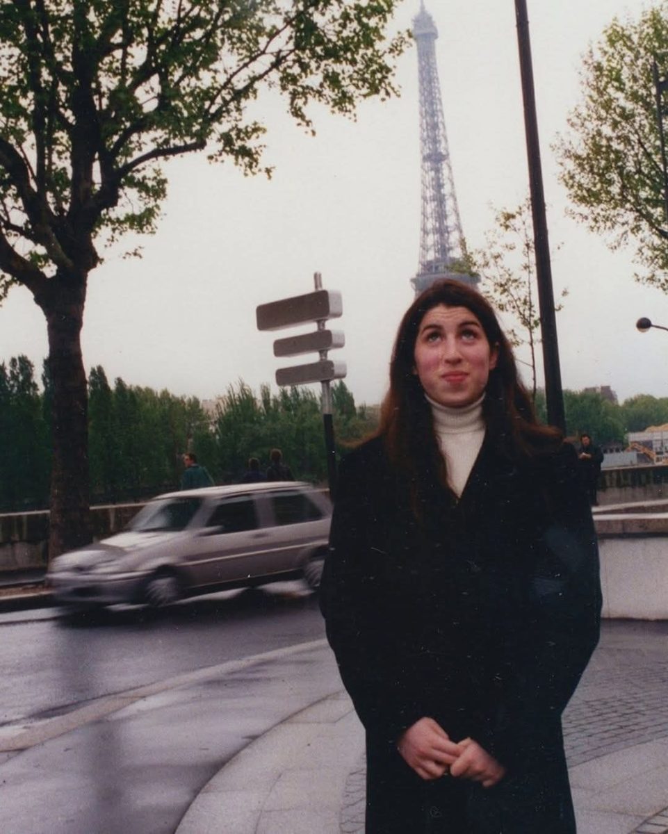 Žena v čiernom kabáte stojí na parížskej ulici s Eiffelovou vežou v pozadí.