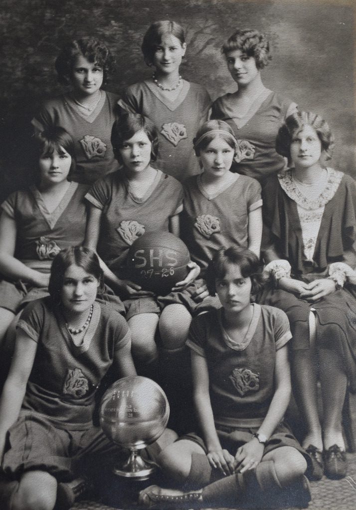 Retro ženský basketbalový tím v uniformách, drží trofej a basketbalovú loptu, ročník 1927-1928.
