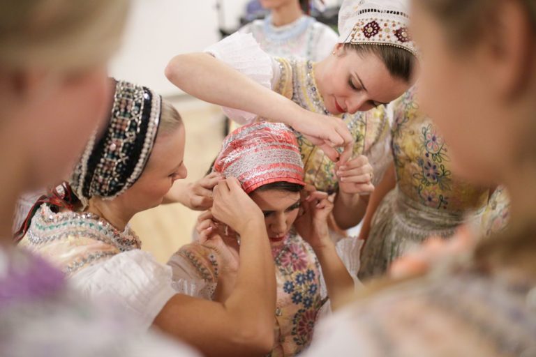 Ženy oblečené v tradičných slovenských krojoch upravujú šatku jednej z nich počas prípravy na folklórnu udalosť.