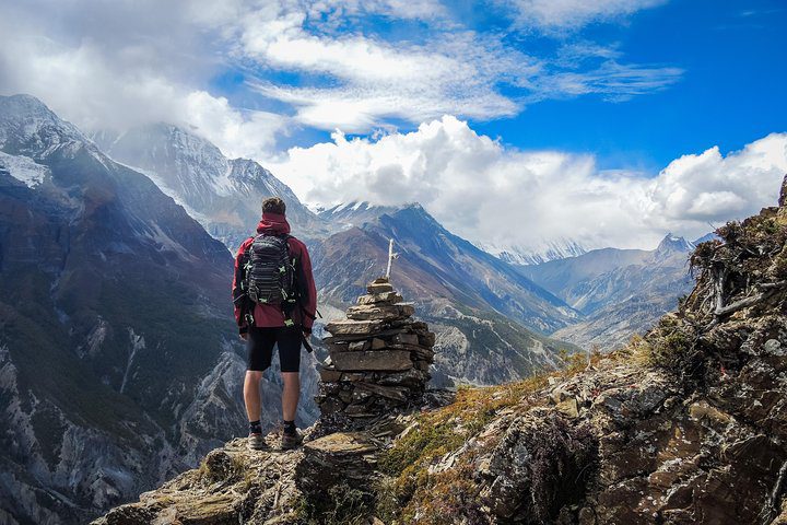 Turista s ruksakom stojí pri kamennom mojejníku a obdivuje pohľad na zasnežené hory pod modrou oblohou.