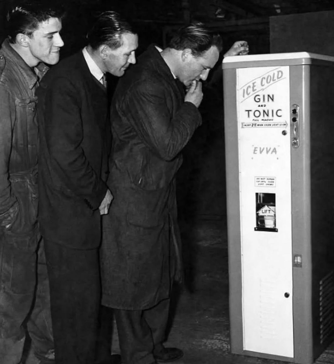 Muži skúmajú starožitný automat na gin s tonikom, zvedaví na jeho funkčnosť v historickom prostredí.
