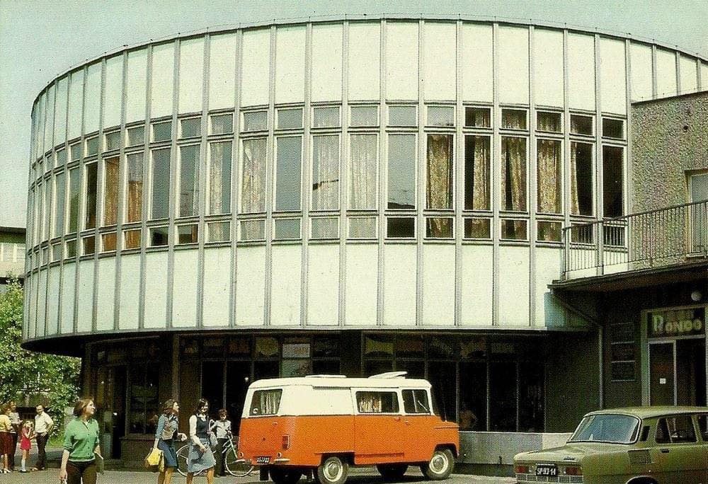 Moderná budova s presklenou fasádou a oranžovou dodávkou, okolo idú chodci.
