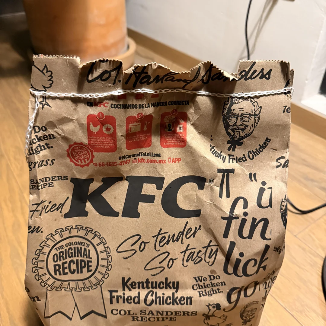 Vrecko od KFC s logom a textom „Kentucky Fried Chicken“ položené na drevenej podlahe.