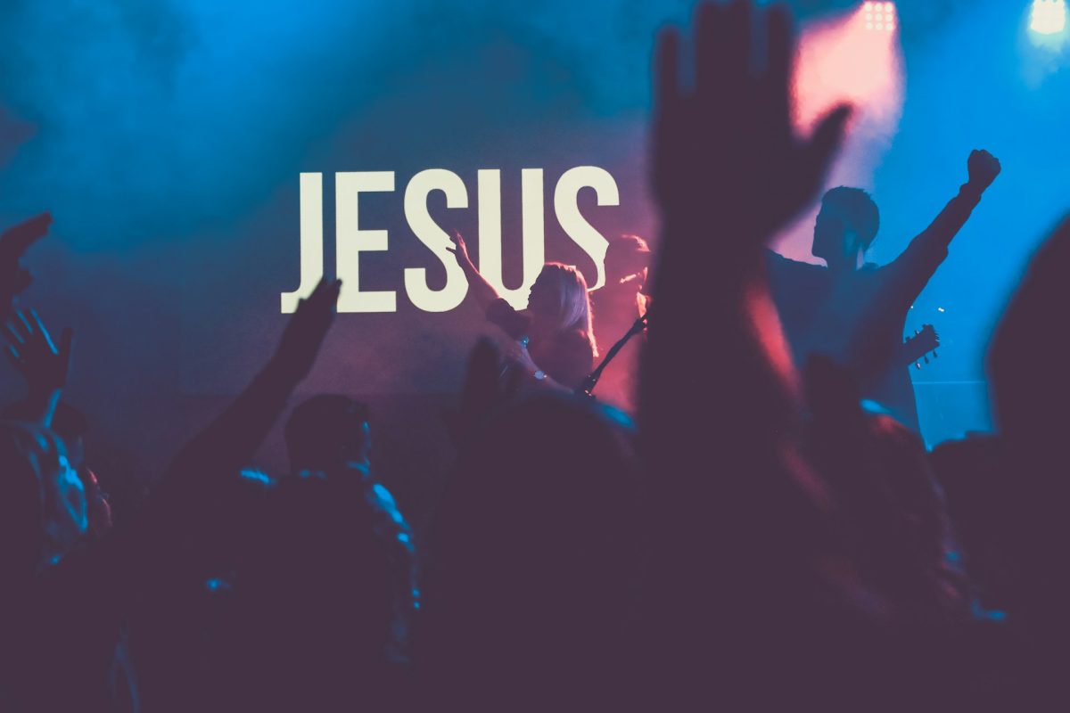 Koncert s nápisom JESUS v pozadí, dav s nadšením zdvíha ruky vo farebnej atmosfére.