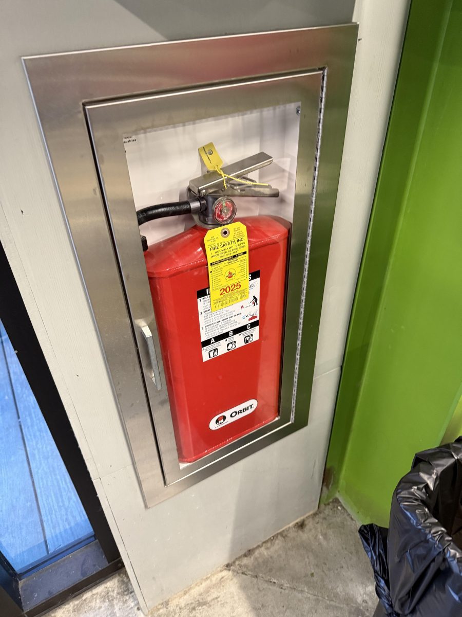 Požiarny hydrant s identifikačnými štítkami v kovovej skrinke na stene vedľa zelenej steny a odpadkového koša.