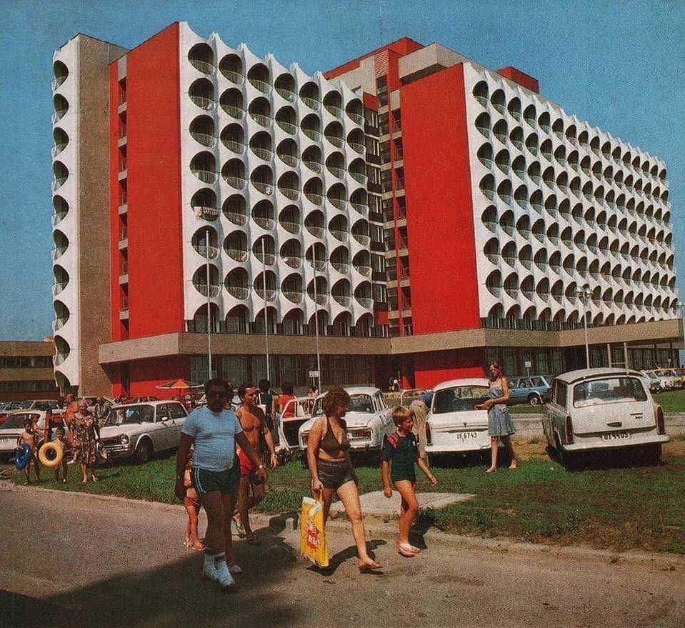 Moderná budova s geometrickými prvkami, ľudia prechádzajúci popri socialistickej architektúre a autách 70. rokov.
