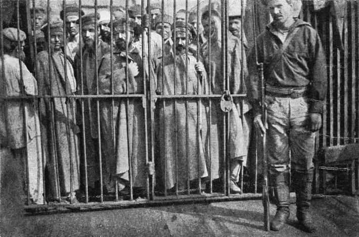 Väzni za mrežami a strážca s puškou v uniformovanom odeve; historická scéna z neznámeho väzenia.