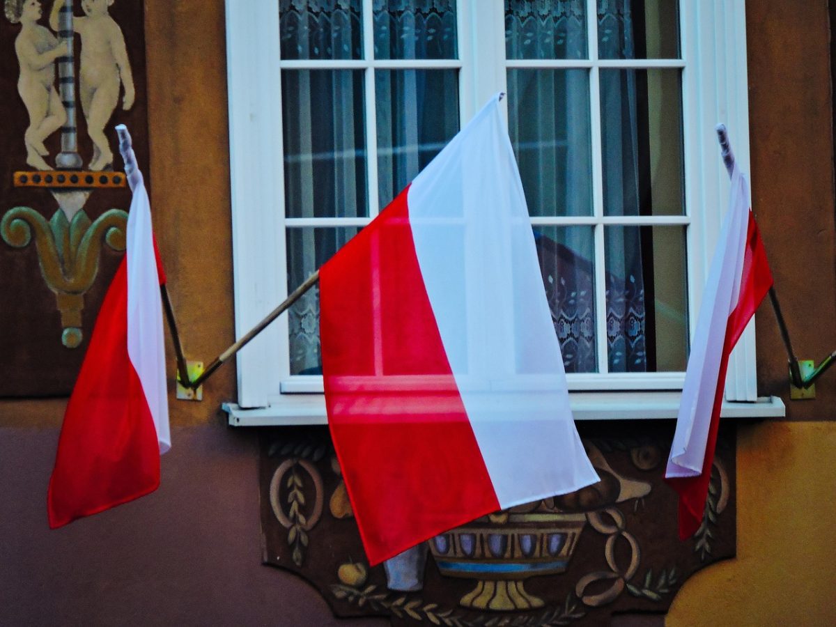 Poľské vlajky visia na historickej budove s výzdobou, zobrazené vedľa zdobenej fasády.