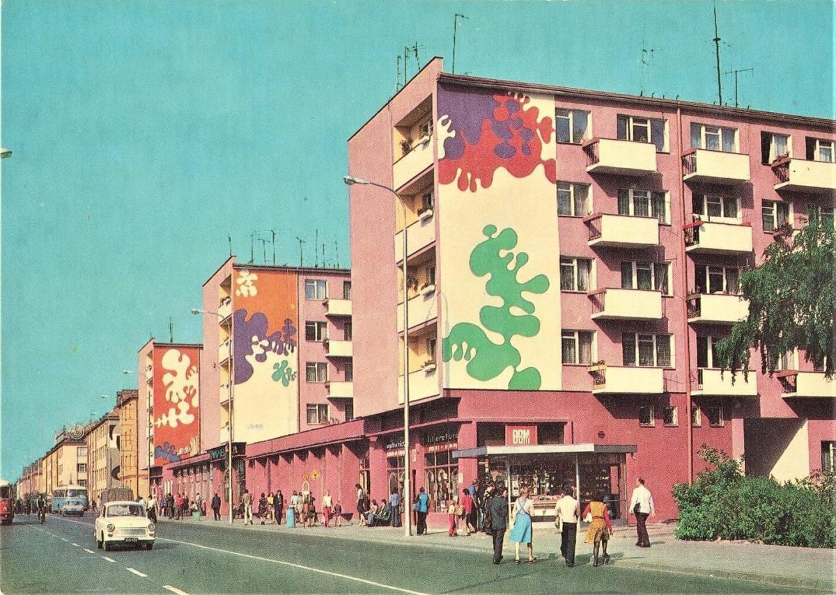 Moderné paneláky s farebnými fasádami a obchodmi, ľudia kráčajúci po rušnej ulici.