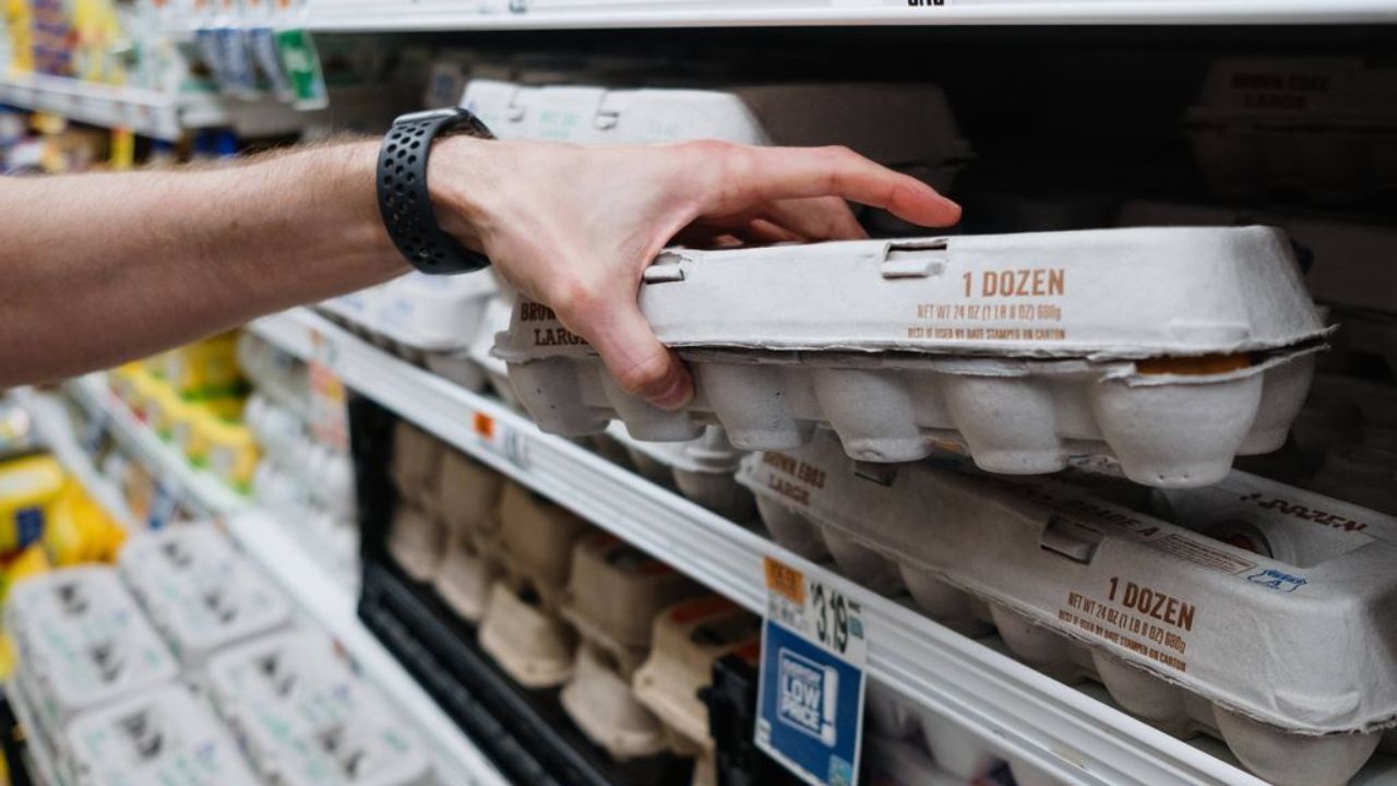 Ruka s čiernym náramkom vyberá balenie vajec z regálu v supermarkete.