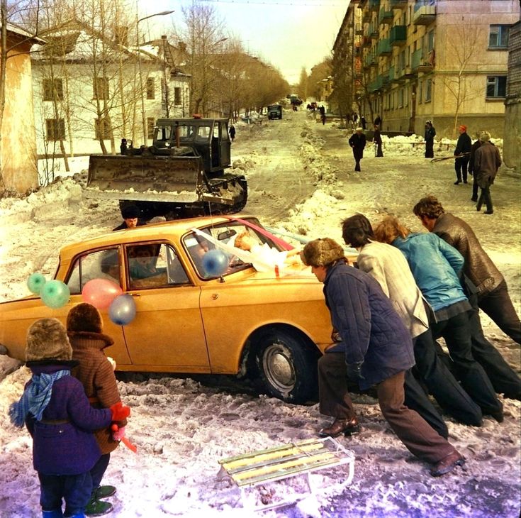 Skupina ľudí tlačí auto na zasneženej ceste, zatiaľ čo buldozér odpratáva sneh v pozadí.