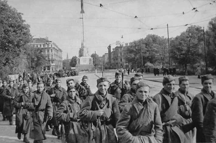 Historická fotografia vojakov pochodujúcich mestom, v pozadí sa týči pamätník a sú vidieť mestské budovy a stromy.