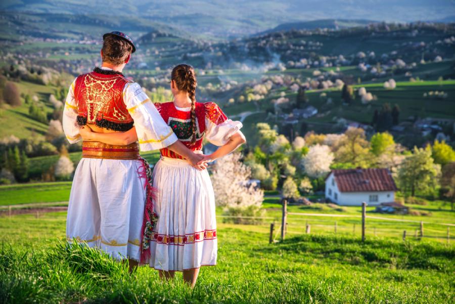 Pár v tradičnom kroji obdivuje malebnú krajinu vidieka s rozkvitnutými stromami a údolím na jar.