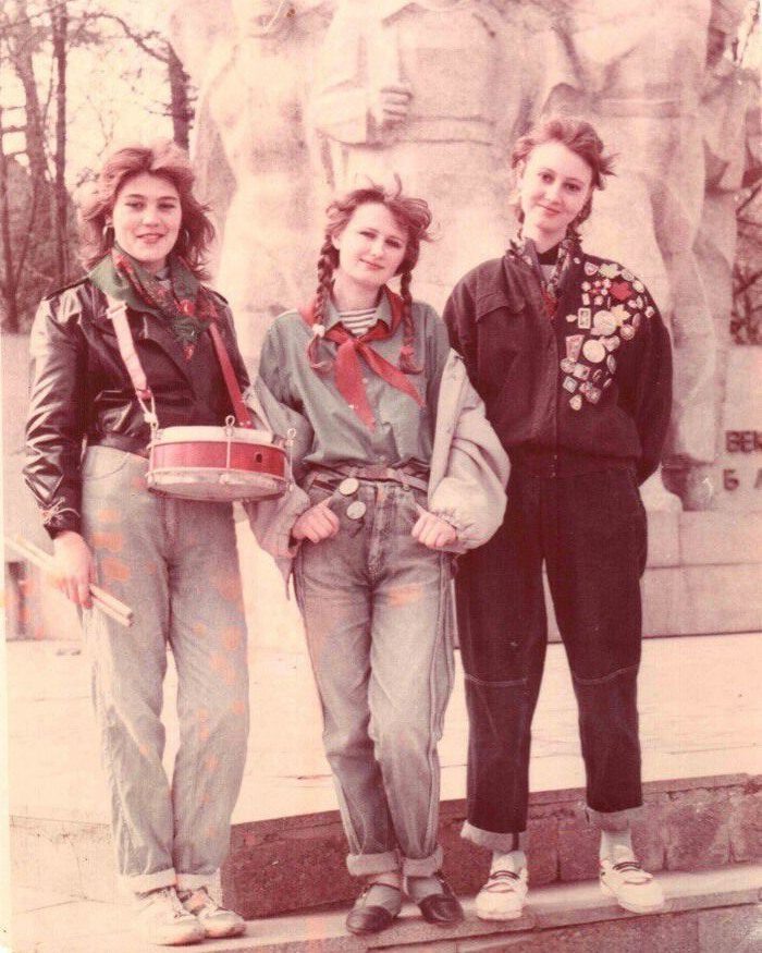 Tri mladé ženy v retro oblečení s bubnom a odznakmi pózujú pred sochou.