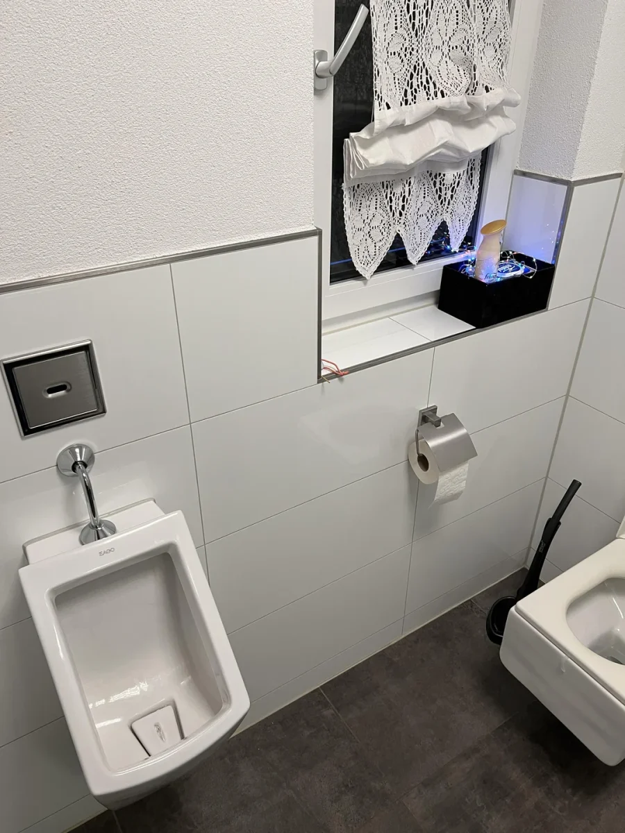 Interiér modernej bielej kúpeľne s pisoárom, toaletou, držiakom na papier a oknom s dekoratívnou záclonou.