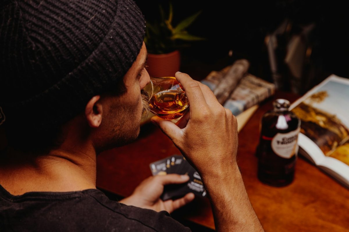 Muž s čiapkou pije whisky pri stole s knihou, kartami a fľašou, domáca relaxácia.