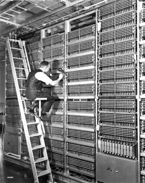Technik opravuje veľký starý telekomunikačný systém, sedí na rebríku a používa nástroje. Retro technológia.