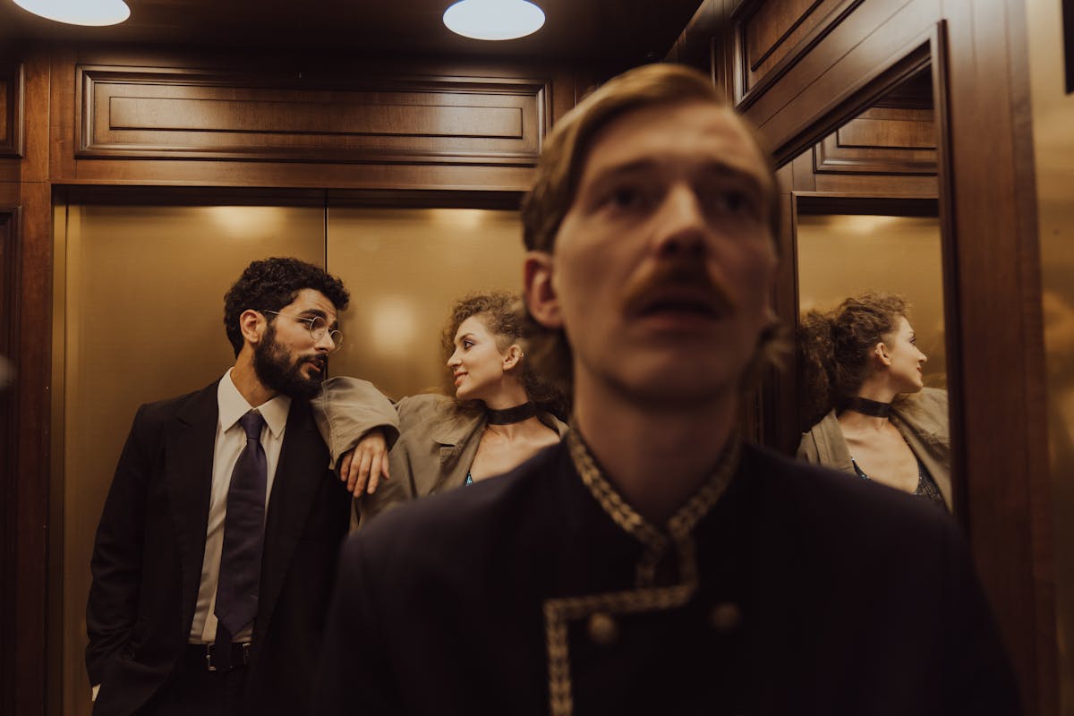 A stylish couple exchanges glances in a wood-paneled elevator alongside a concierge.
