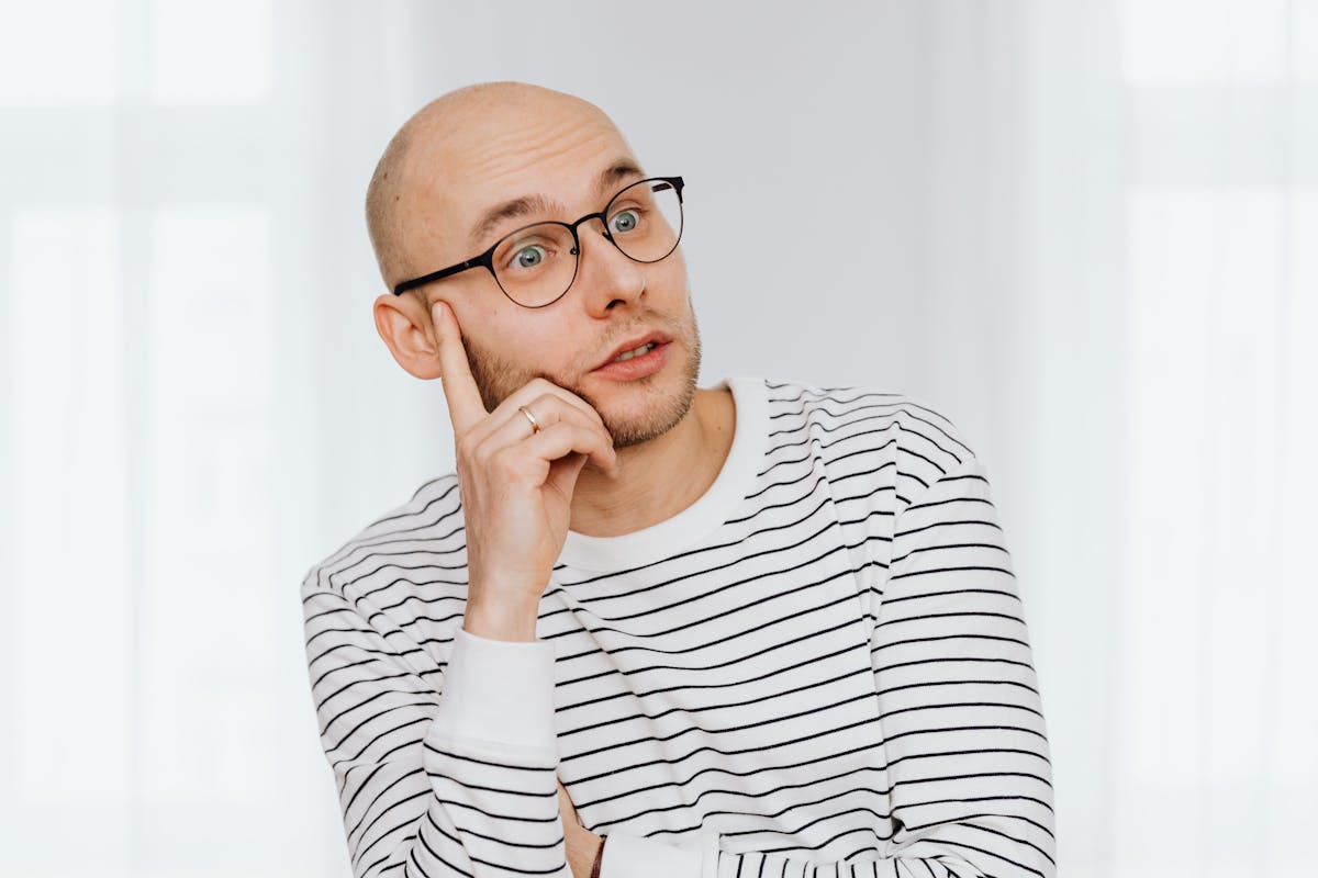 A bald man wearing glasses and a striped shirt gazes thoughtfully indoors.
